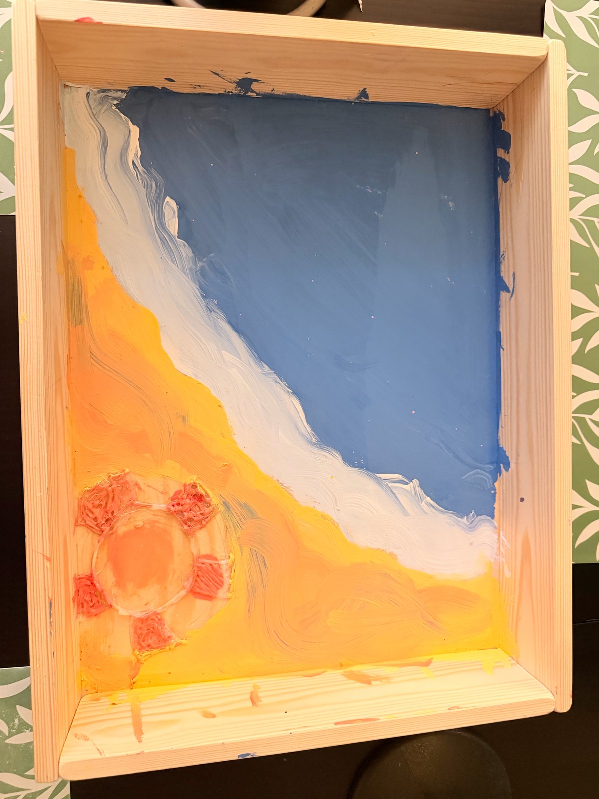 A painting in a wooden tray: blue sky, a white wave sweeping diagonally, orange and yellow below, and a sun with a pink-red outline in the bottom-left corner.