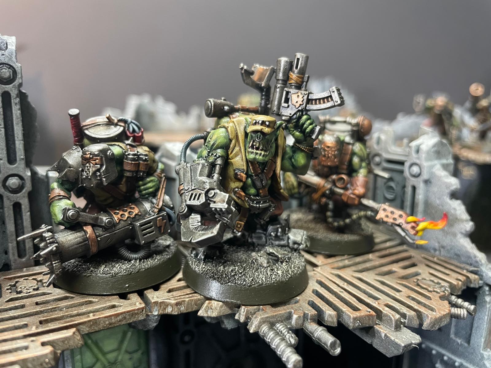 A close-up of three painted orc miniatures on industrial terrain, one with a mechanical claw.
